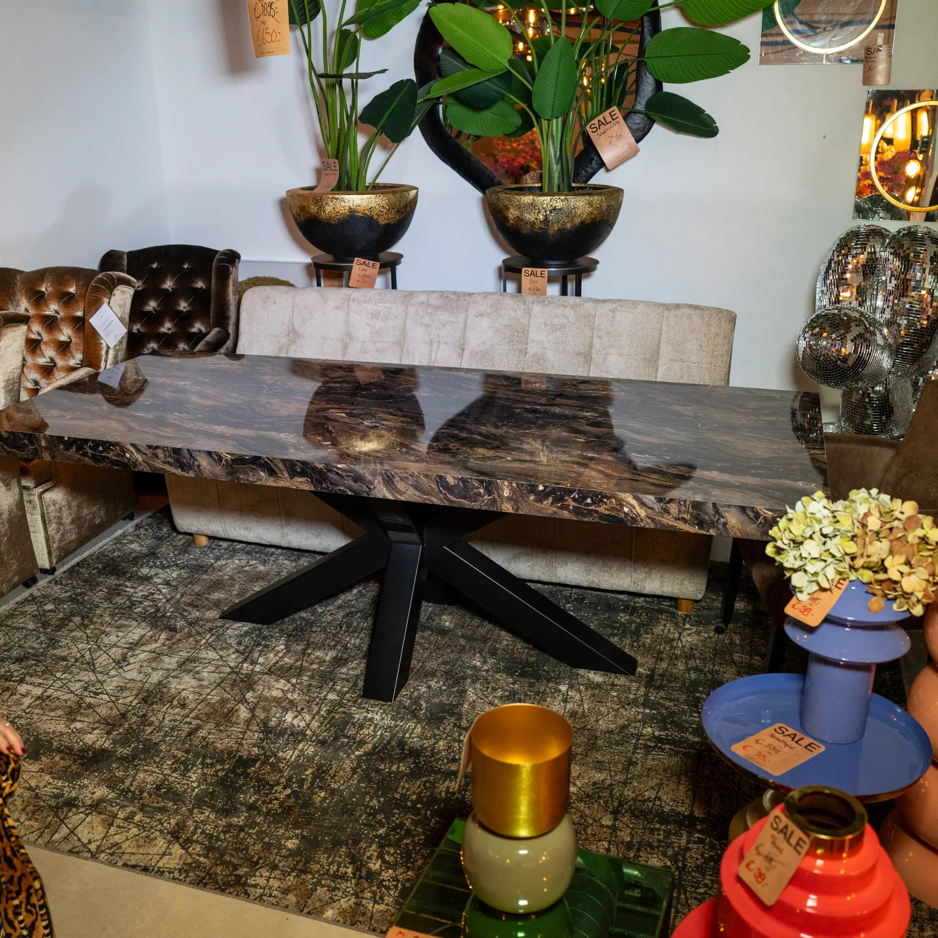 Showroom scene with a marble table, tufted leather seats, tall potted plants, and colorful stacked vases labeled SALE on tags to the right.