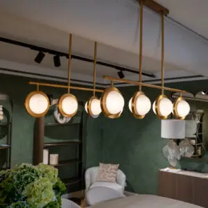 Gold circular pendant light fixture hanging over a green textured wall with seating and plants in a showroom.