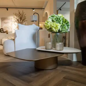 Living room showroom scene with a round dark coffee table and a glass vase of green hydrangeas in the foreground, soft seating and decorative lamps in the background.
