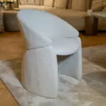 Close-up of a light gray textured fabric chair with a curved back in a living room setting.