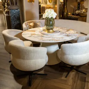 Round marble dining table surrounded by beige upholstered chairs with a glass vase of white roses as the centerpiece in a warm, stylish room.