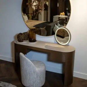 Round illuminated vanity mirror mounted on a light wall, reflecting a bedroom with a bed, pillows, and decor.