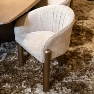 Close-up of a beige upholstered chair with dark brown legs tucked under a light wood table on a shaggy brown carpet.