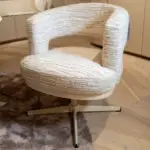 Close-up of a beige, textured, circular chair with layered fabric upholstery and metal legs on a wooden floor.
