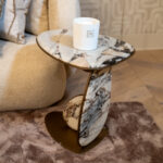 White cylindrical candle in a holder with a square logo sits on a round marble table in a neutral-toned room.