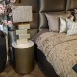 Luxurious bedroom corner with a crystal lampshade on a gold stacked pedestal beside a tufted headboard and beige quilted bedding.
