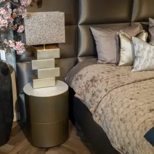 Luxurious bedroom corner with a crystal lampshade on a gold stacked pedestal beside a tufted headboard and beige quilted bedding.
