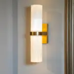 White cylindrical wall-mounted lamp with warm glow and brass mount, against a gray wall with a yellow backing panel.