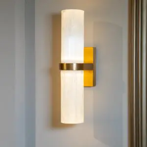 White cylindrical wall-mounted lamp with warm glow and brass mount, against a gray wall with a yellow backing panel.
