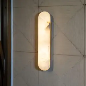 Vertical frosted glass wall sconce casting warm light on a textured wall with stacked chevron patterns.