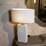 A close-up of a woven fabric lampshade atop a wooden pedestal lamp on a shelf, with a patterned tiled wall in the background.