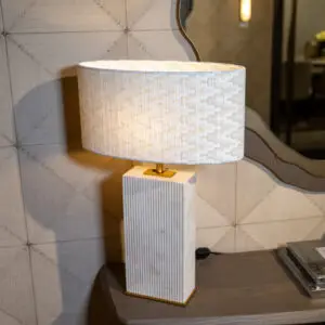 A close-up of a woven fabric lampshade atop a wooden pedestal lamp on a shelf, with a patterned tiled wall in the background.