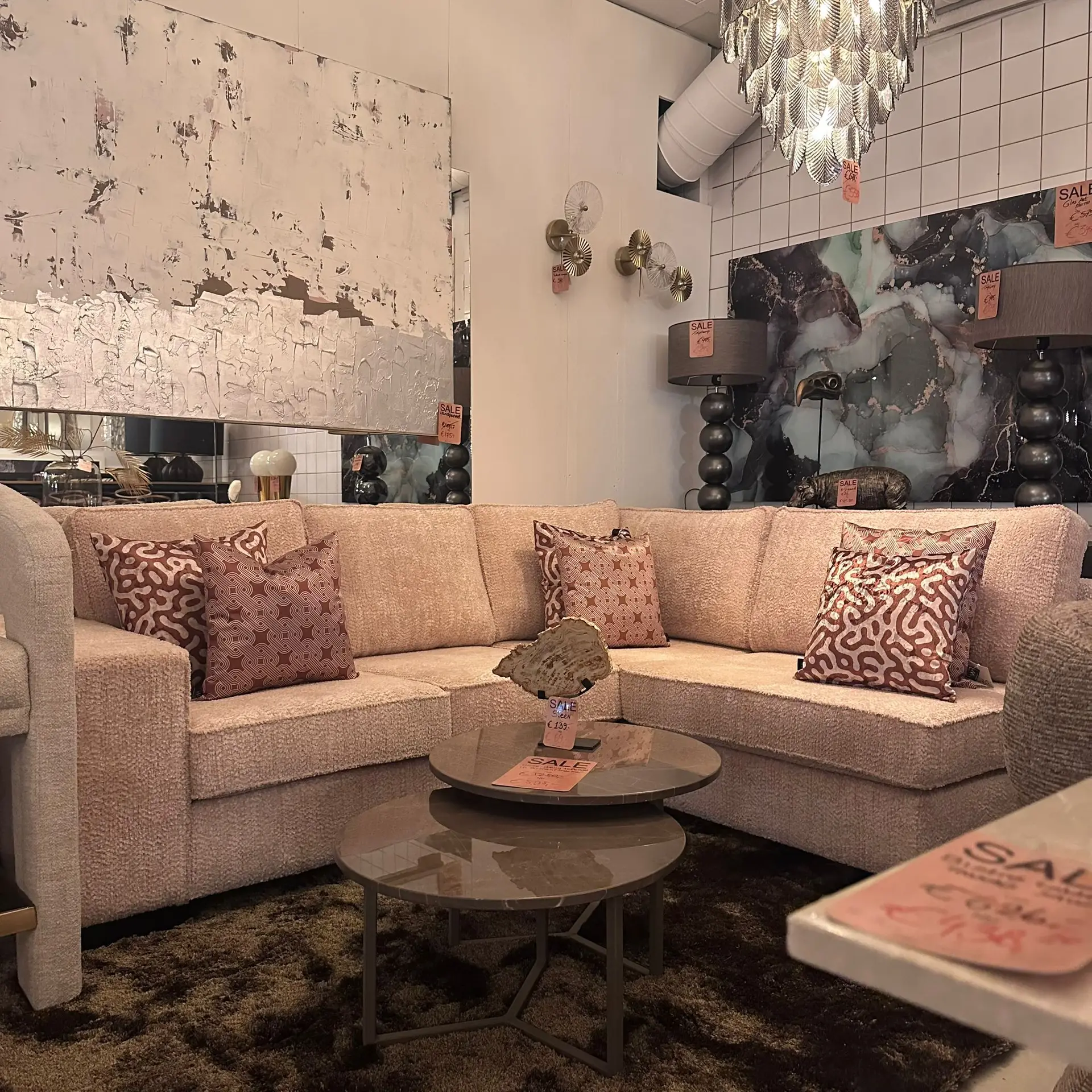 Beige sectional sofa with brown patterned pillows in a furniture showroom, with glass coffee tables and sale tags.