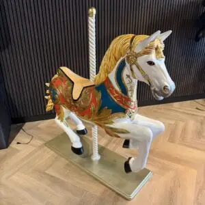 Carousel horse sculpture with a gold mane, ornate blue and red saddle, indoors on a metal base with a striped wall behind it.
