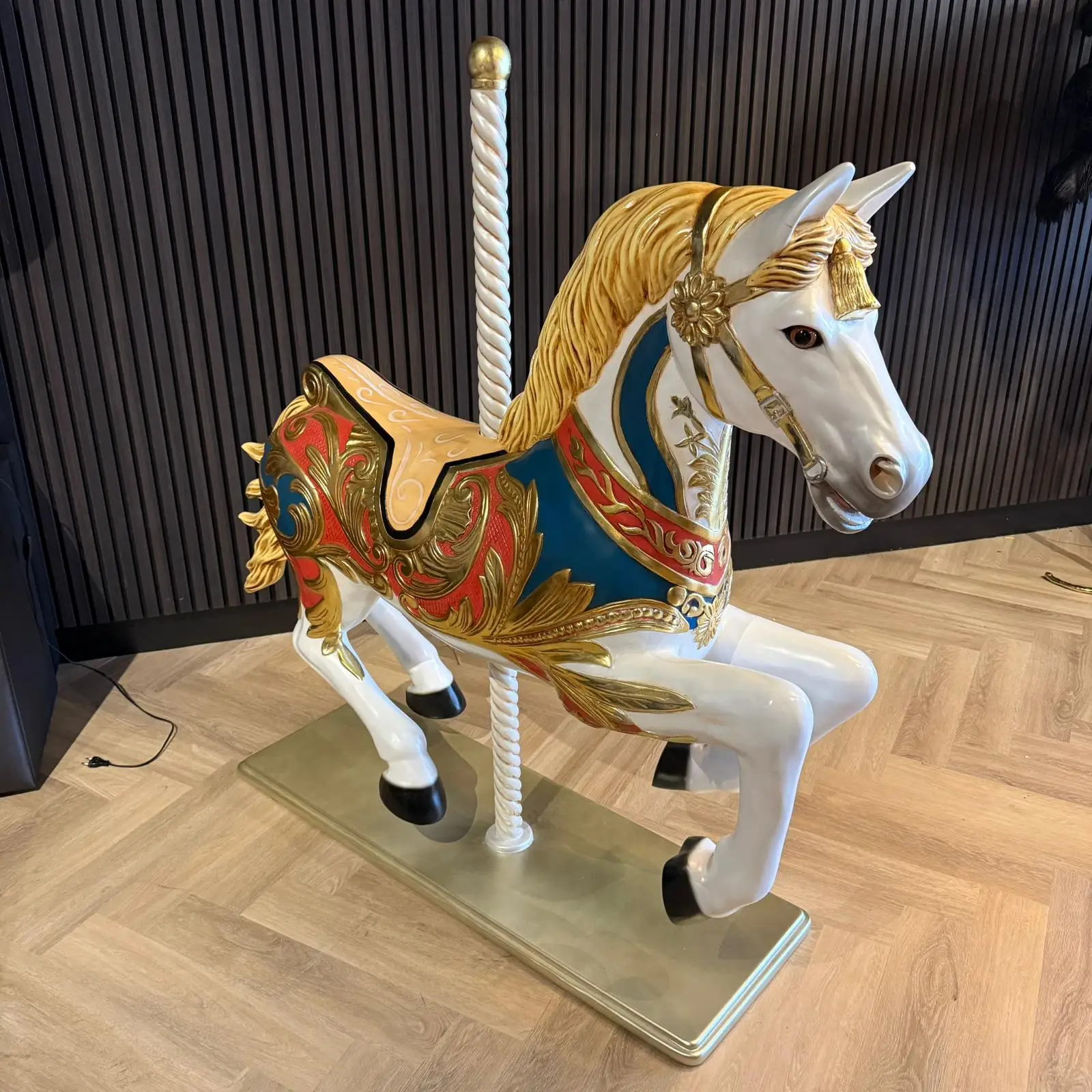 Carousel horse sculpture with a gold mane, ornate blue and red saddle, indoors on a metal base with a striped wall behind it.