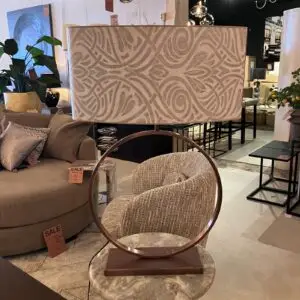 Patterned lampshade on a round metal stand atop a marble table in a furniture showroom, with a beige sofa and sale tags in view.
