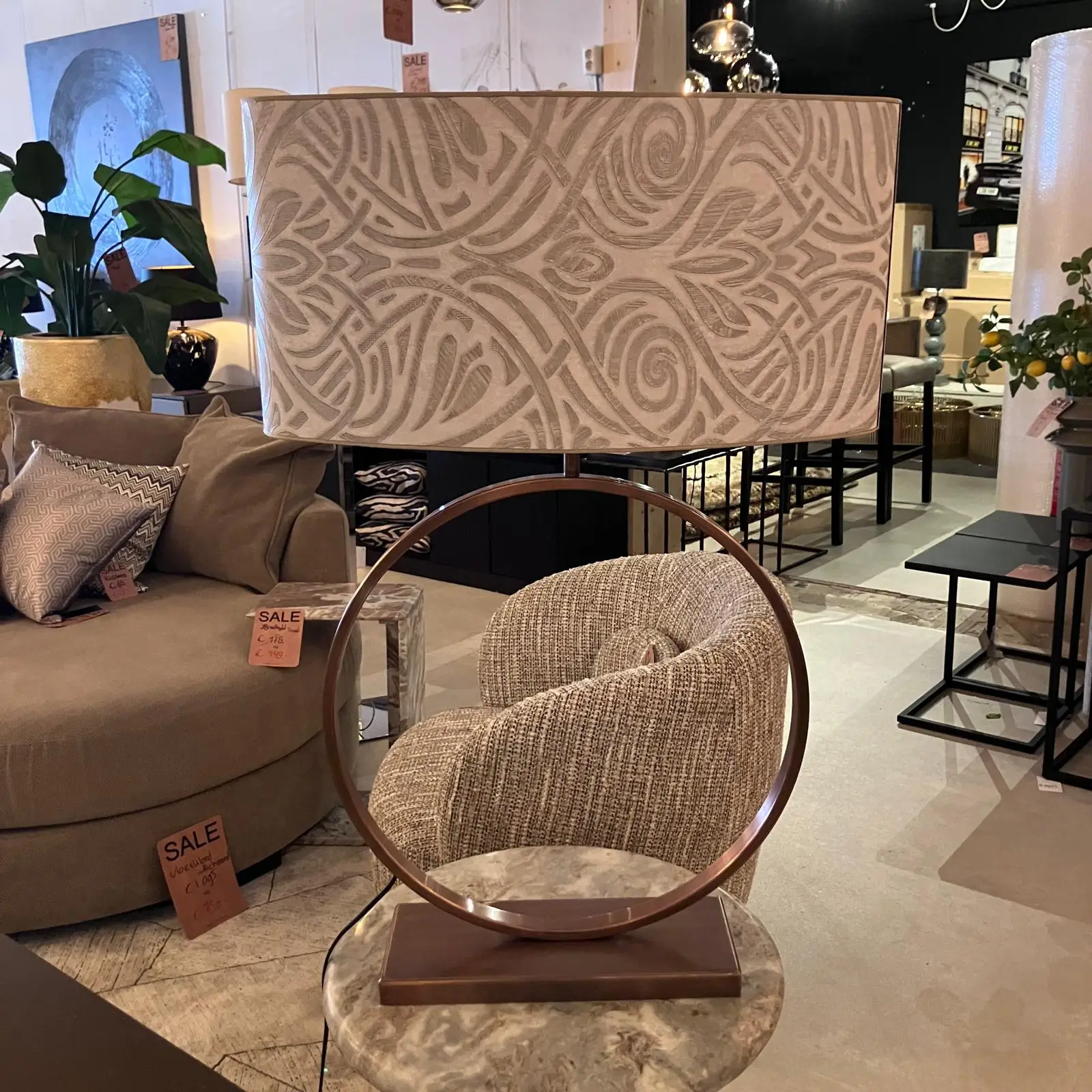 Patterned lampshade on a round metal stand atop a marble table in a furniture showroom, with a beige sofa and sale tags in view.