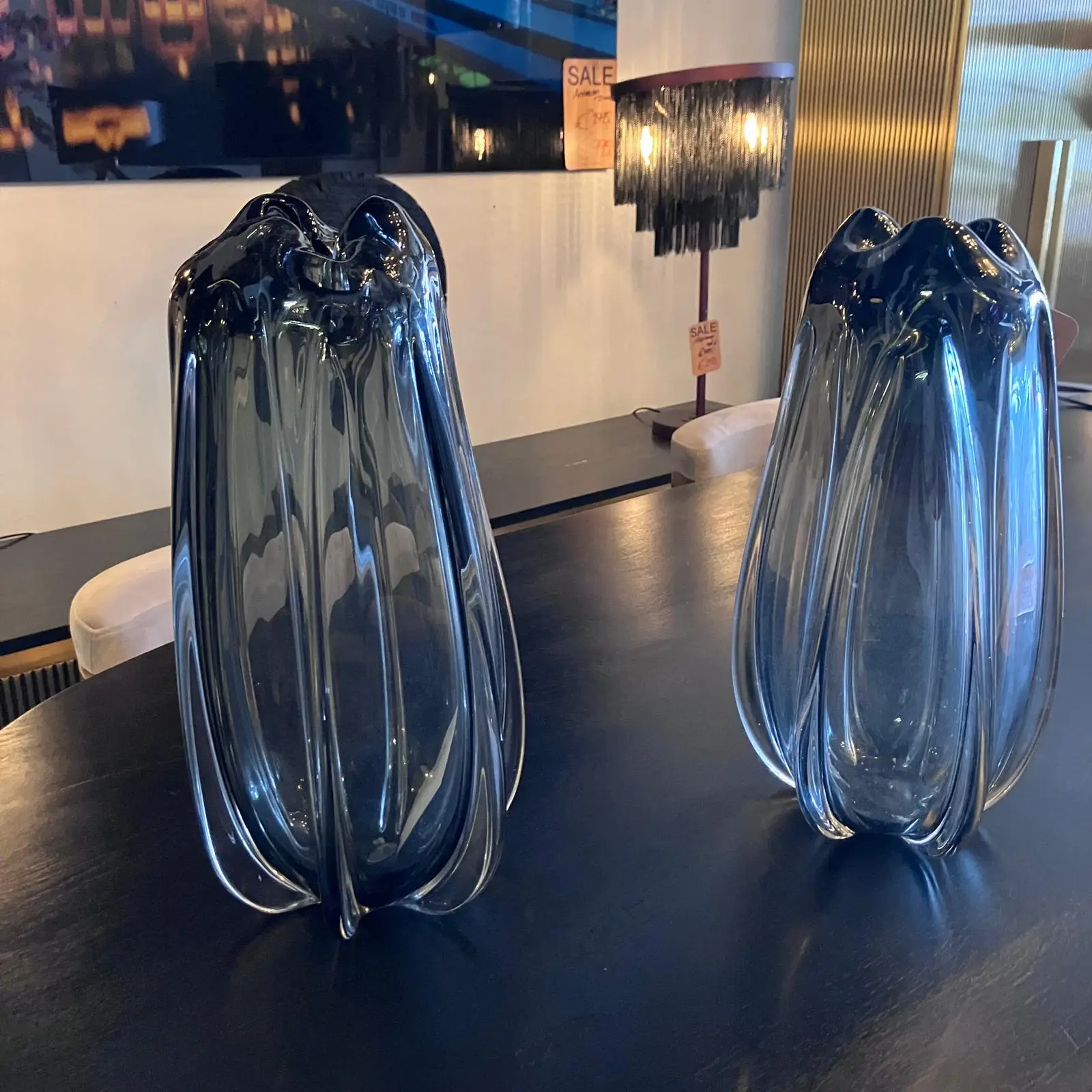 Two tall, curved blue glass sculptures sit on a dark table in a modern showroom, with warm-lit lamps in the background.
