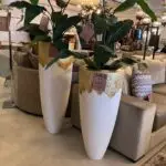 Indoor furniture showroom; tall white planters with gold accents, green plants, and beige sofas with sale tags