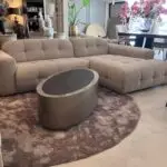 Light beige tufted sectional sofa on a circular brown rug with a silver metal coffee table in a showroom setting with plants in the background and pink flowers on a table above.