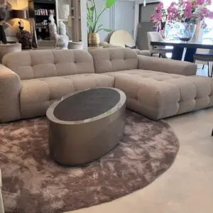 Light beige tufted sectional sofa on a circular brown rug with a silver metal coffee table in a showroom setting with plants in the background and pink flowers on a table above.