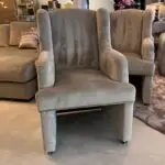 Group of light gray upholstered armchairs on casters in a showroom, with a patterned pillow and other furniture in the background.