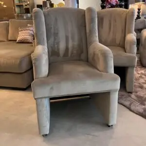 Group of light gray upholstered armchairs on casters in a showroom, with a patterned pillow and other furniture in the background.