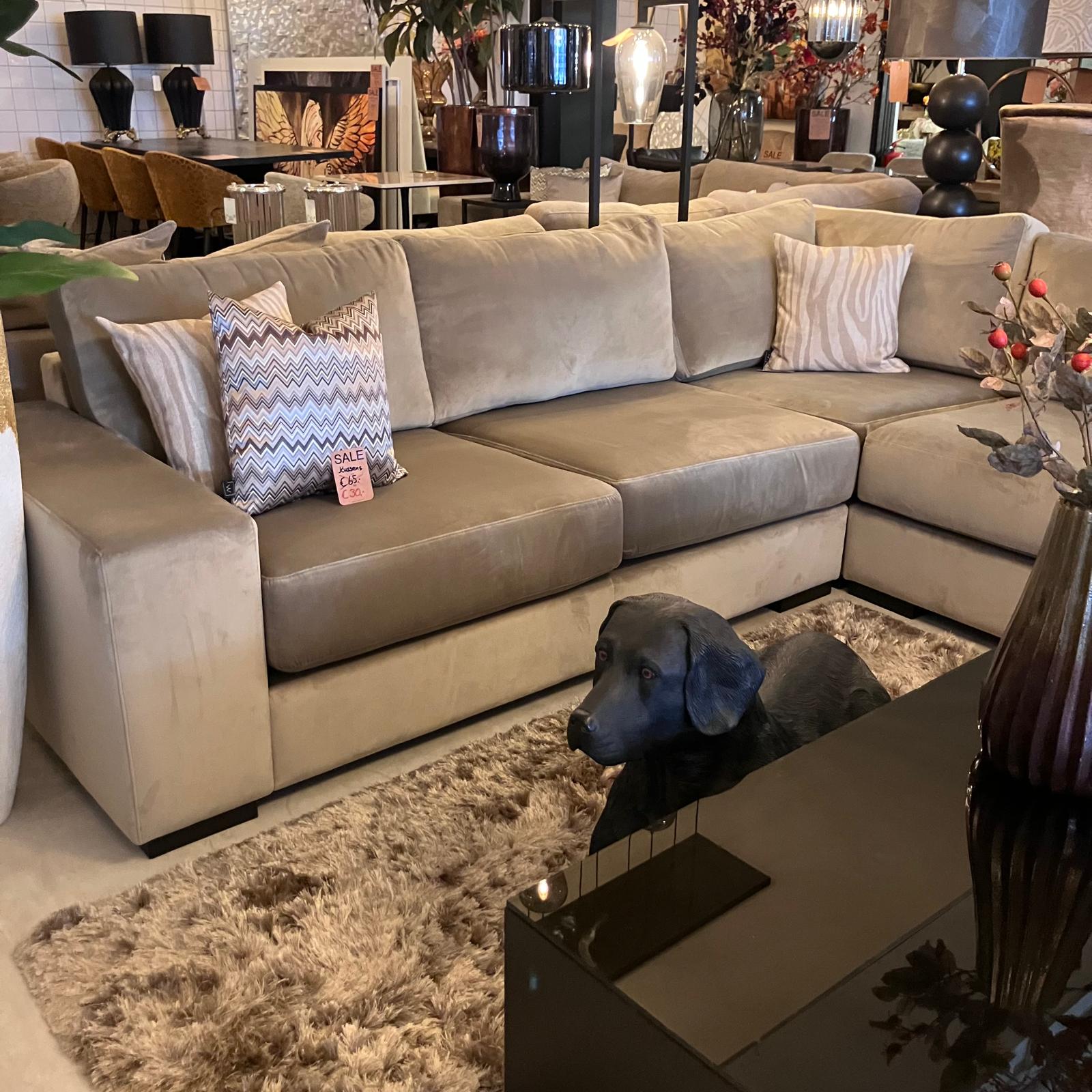 Gray sectional sofa with striped and zigzag throw pillows in a showroom, sale tag visible on one pillow, a black dog sculpture resting on a shaggy beige rug in front of a glass coffee table.