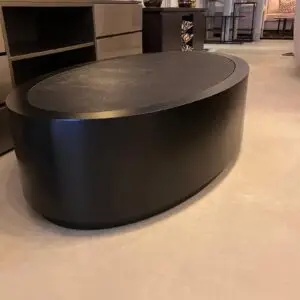Large round black coffee table with a textured top in a furniture showroom, with cabinets and patterned textiles in the background.