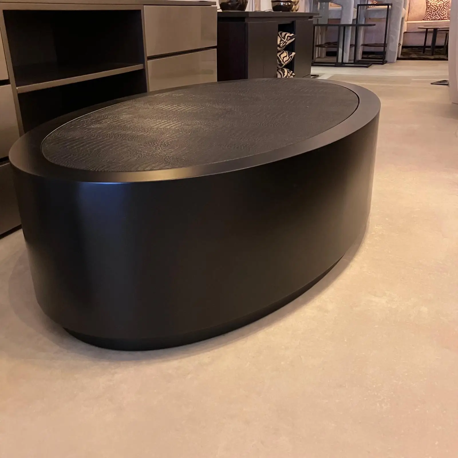 Large round black coffee table with a textured top in a furniture showroom, with cabinets and patterned textiles in the background.