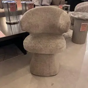 Beige sculptural chair with a thick curved back and pedestal base in a showroom, surrounded by sale-tagged display furniture.