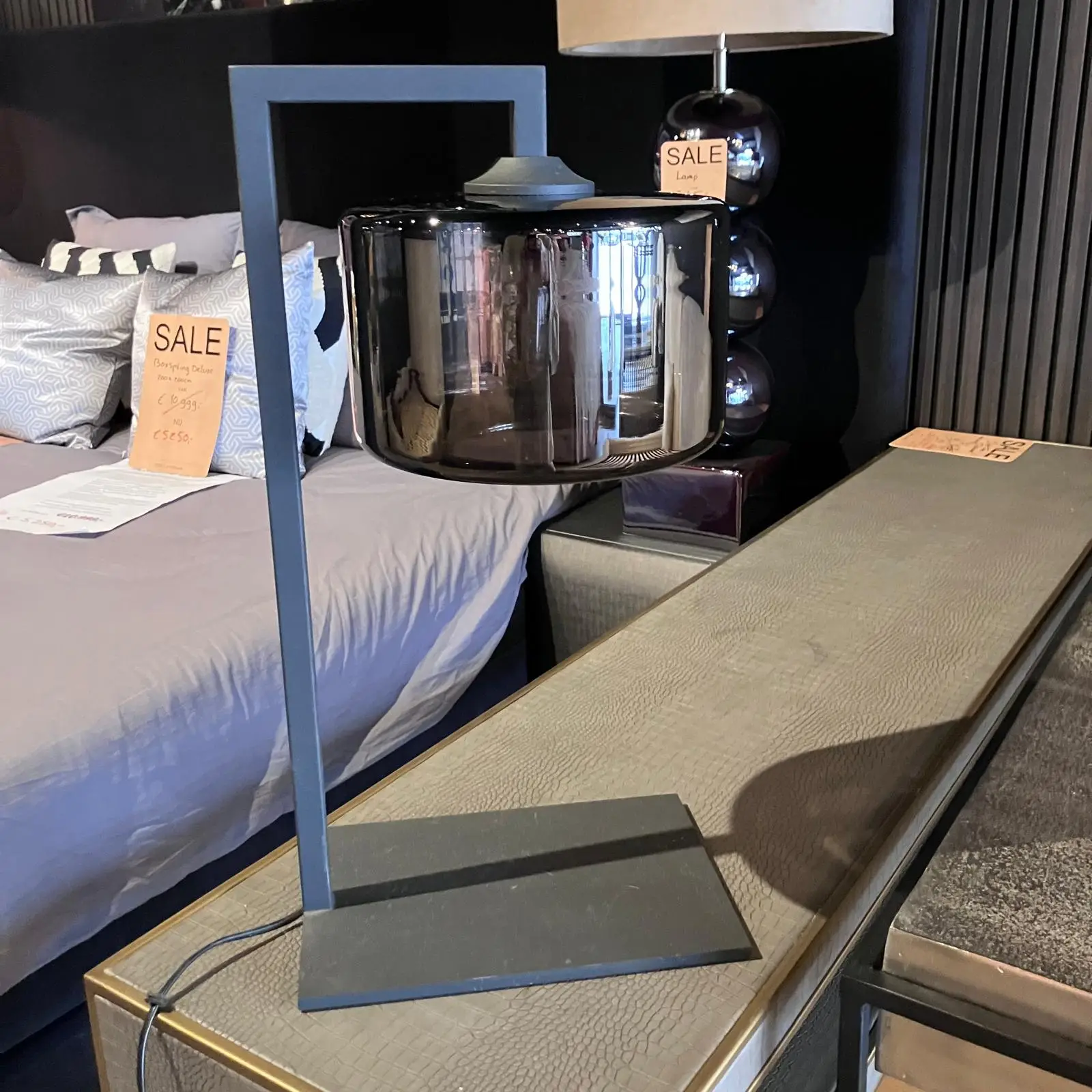 Glossy black glass pendant lamp on a metal stand in a showroom, with a bed and orange sale signs in the background.