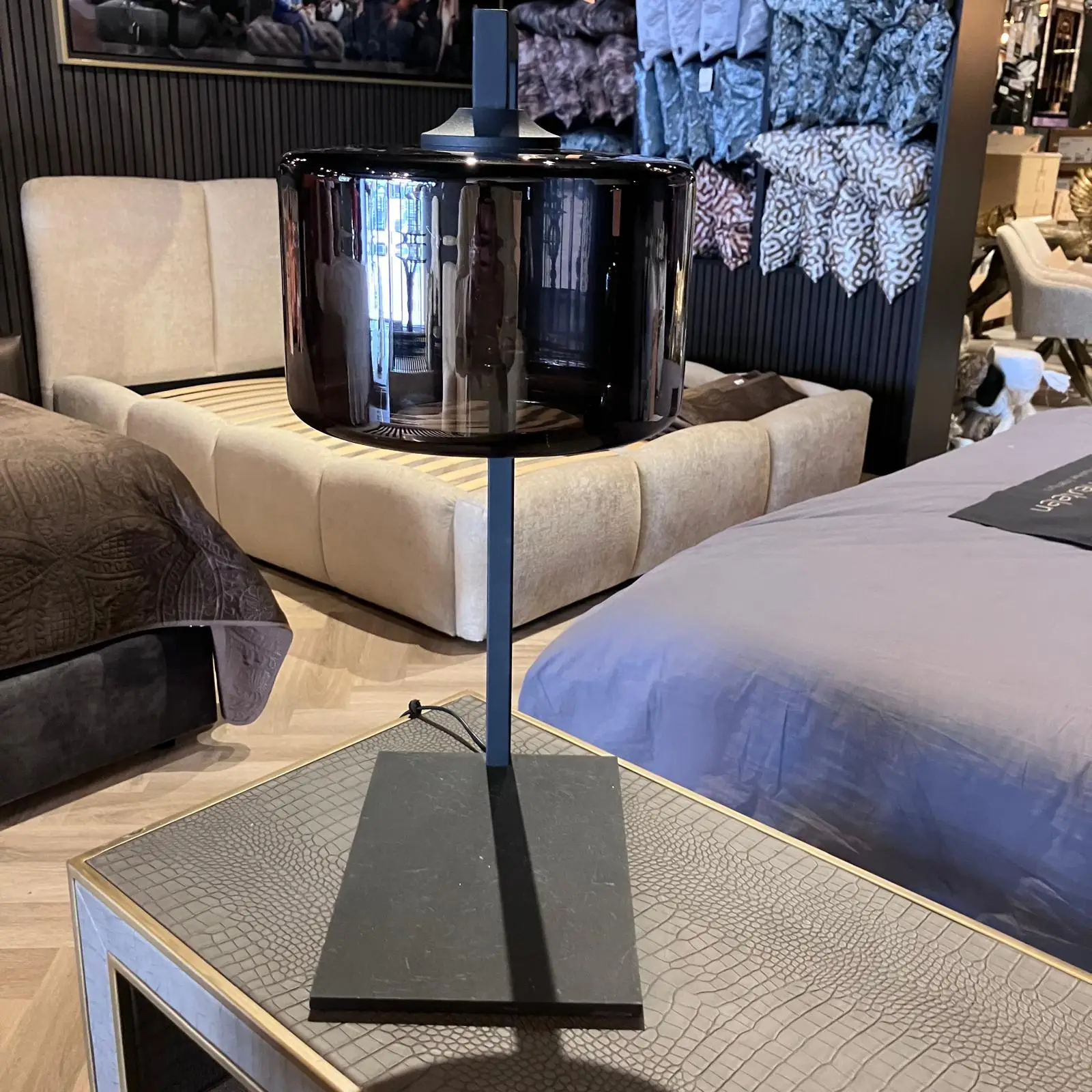 Glossy black cylindrical table lamp on a slim blue stand in a home decor showroom with a bed and seating in the background