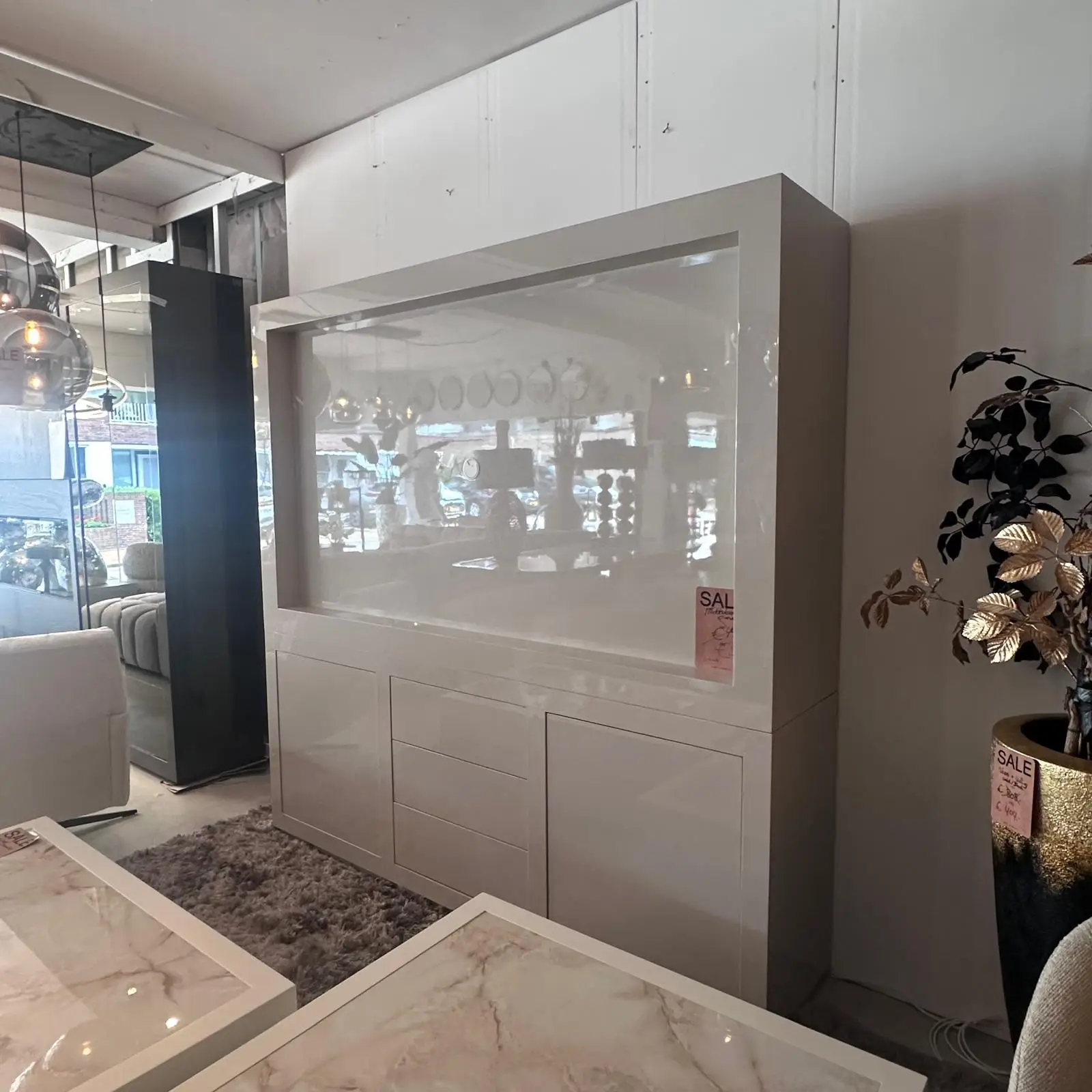 White high-gloss display cabinet with a glass front in a furniture showroom, reflecting the store lights and shelves nearby.