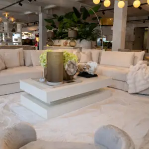 Neutral showroom living room with light beige sectional sofa, plush cushions, and a white shag rug displaying modern decor on a white coffee table.