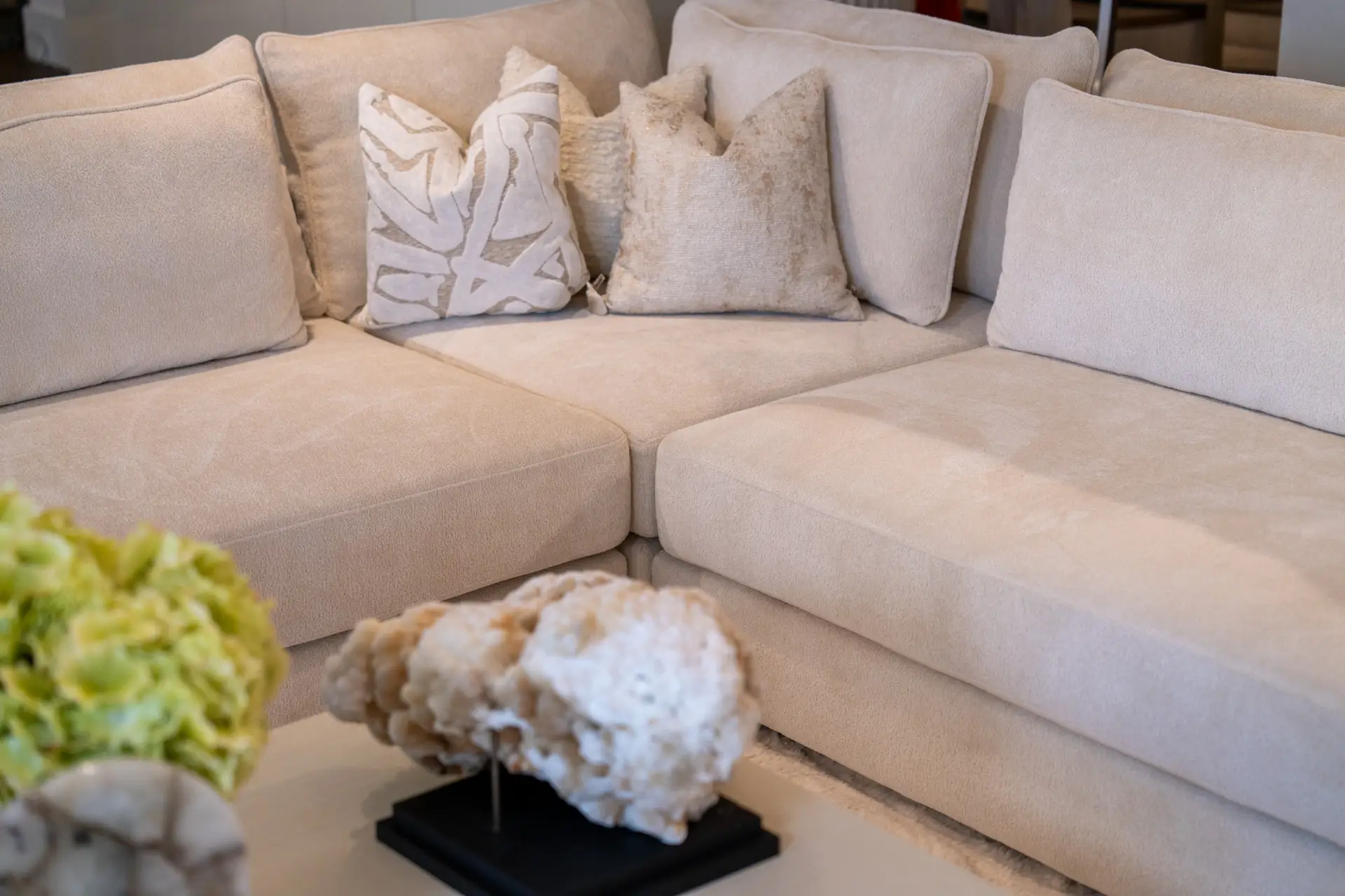 Beige sectional sofa with several light-colored cushions, including a white patterned one, in a cozy living room. Readable and concise for navigation or layout.