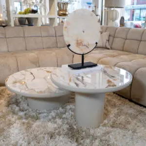Round stone disc sculpture with subtle rust marks on a black stand, placed on a marble coffee table in a light showroom setting.