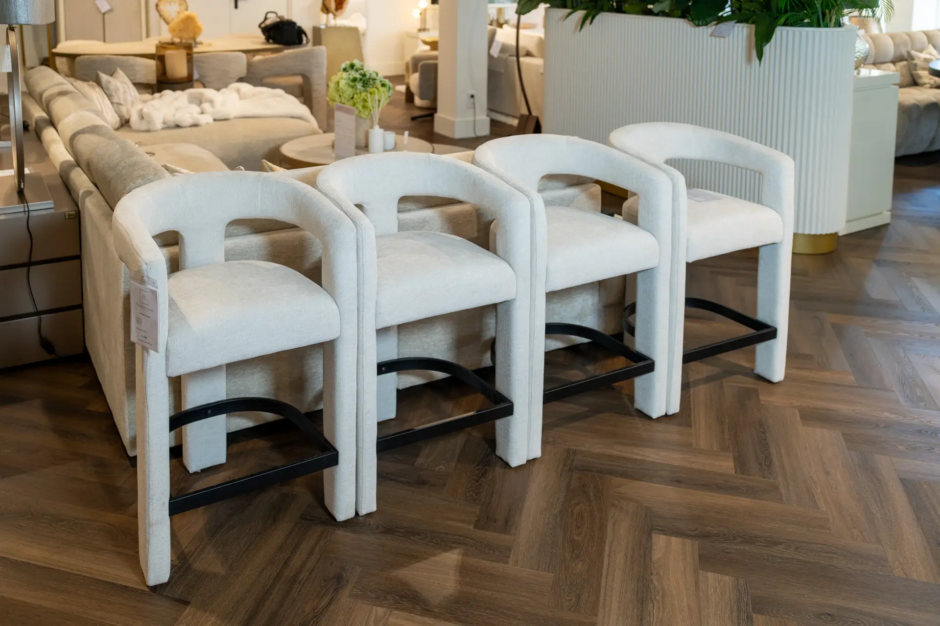 Row of light beige upholstered chairs with black metal bases in a furniture showroom, with sofas and plants in the background.
