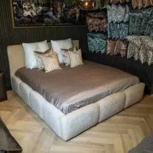 Large beige upholstered bed with cream and patterned pillows in a showroom, wall with a framed collage and fabric swatches to the right.",