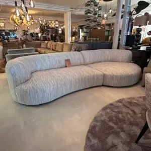 Curved light gray upholstered sofa with a small sale tag on the backrest, showroom filled with other sofas and modern lighting in the background.