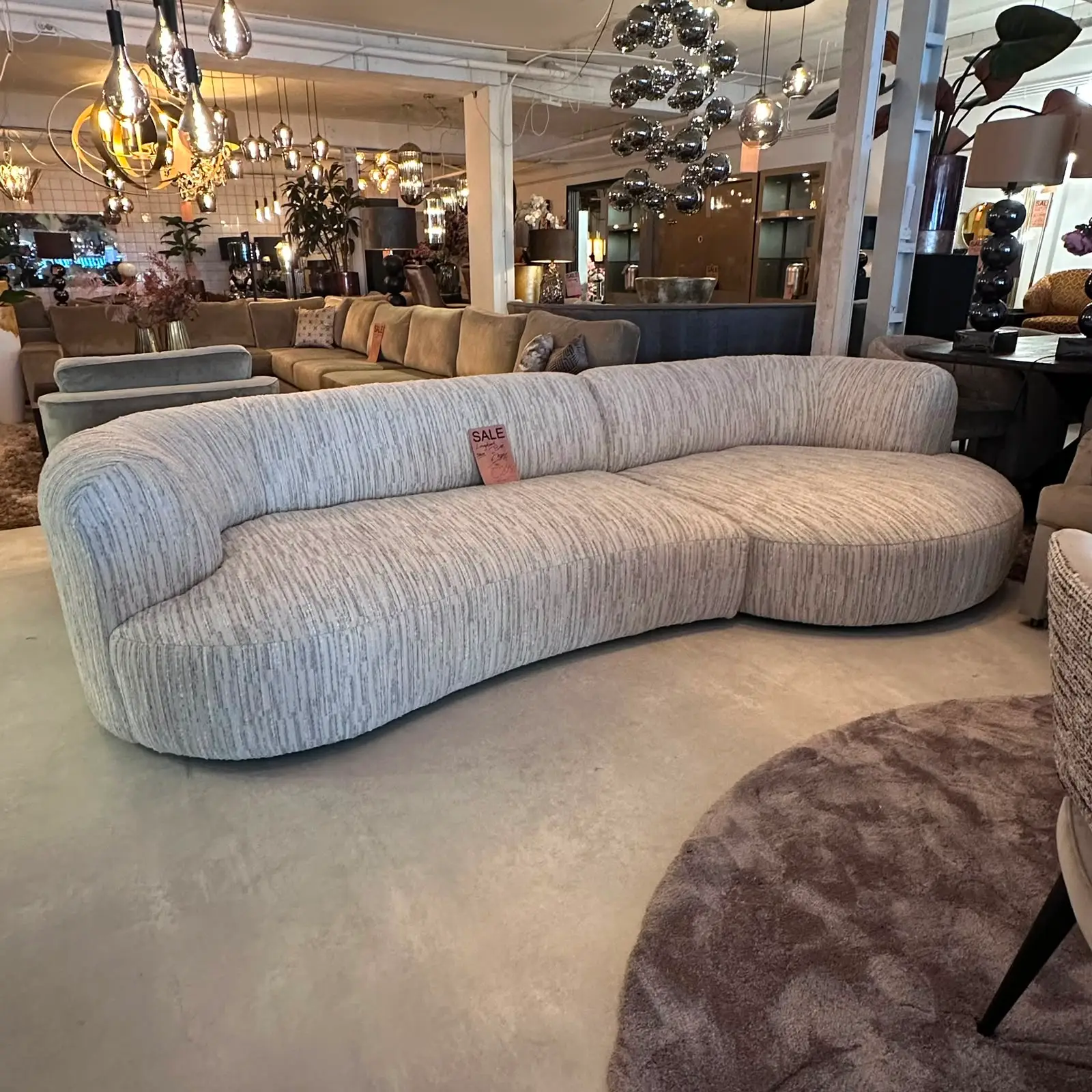Curved light gray upholstered sofa with a small sale tag on the backrest, showroom filled with other sofas and modern lighting in the background.