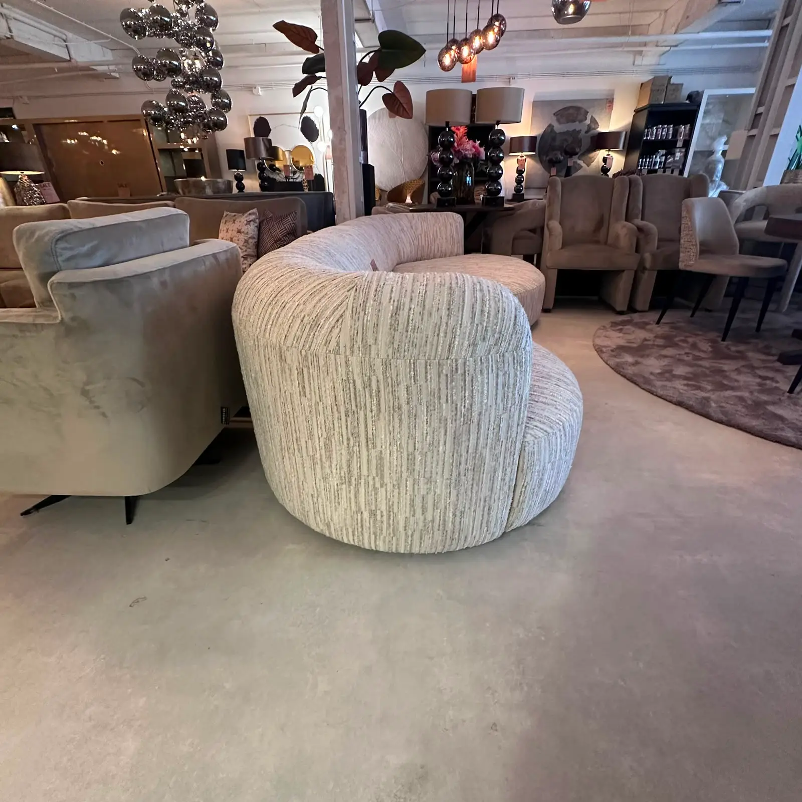 Round beige striped fabric ottoman in a furniture showroom, surrounded by sofas and lounge chairs.