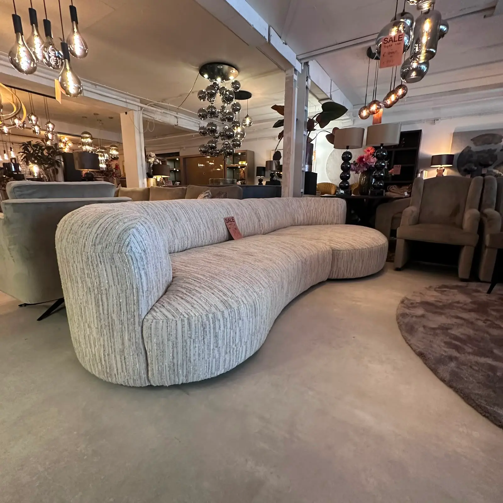 Curved striped light-gray sofa with a sale tag in a furniture showroom, surrounded by other sofas, chairs, lamps and rugs.