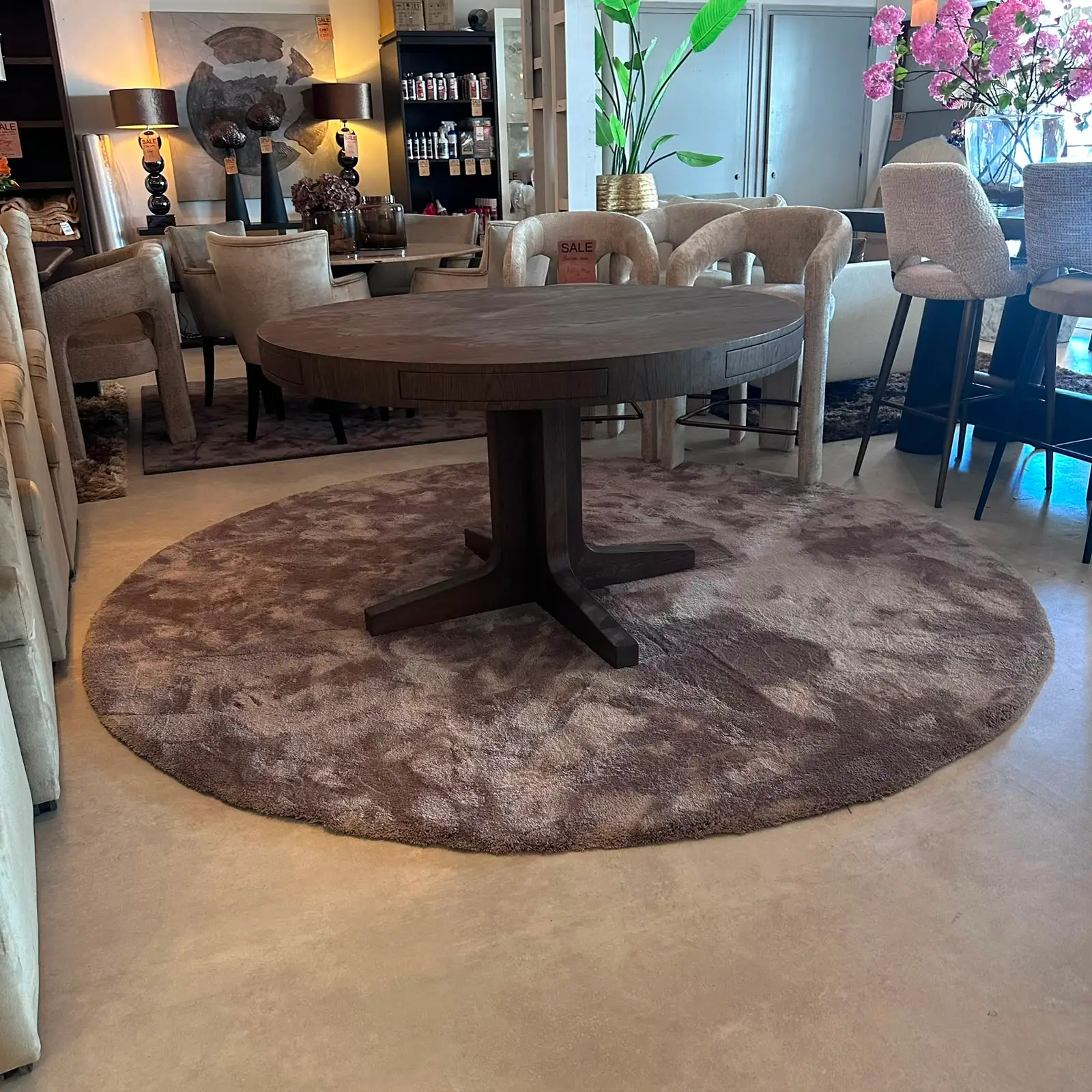 Round wooden pedestal table on a circular brown rug, surrounded by beige upholstered chairs in a furniture showroom.