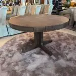 Round dark wood dining table with a pedestal base in a furniture showroom, surrounded by pale upholstered chairs.