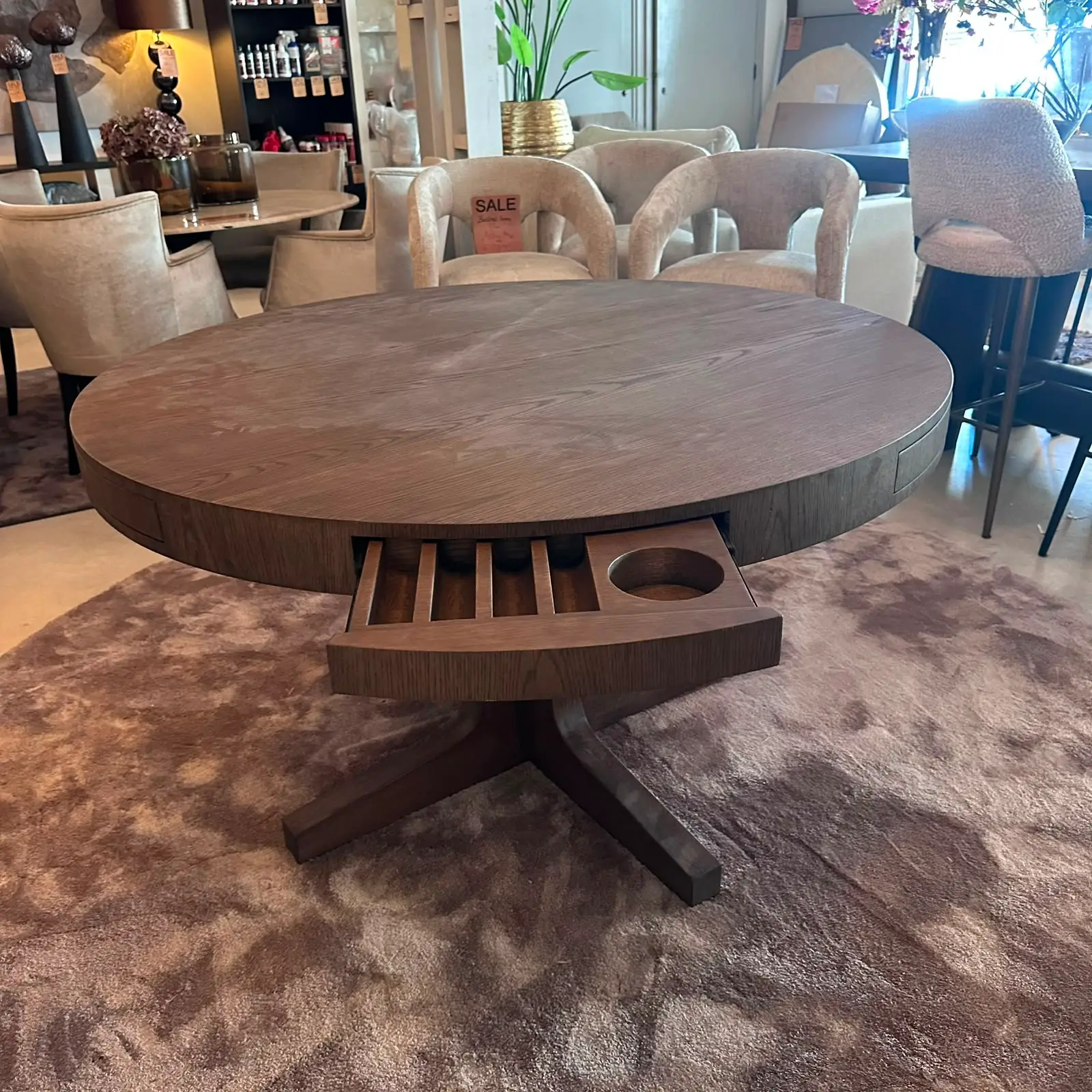 Round dark wood coffee table with built-in cup holders and a slot tray, supported by a cross-shaped pedestal, in a furniture showroom.