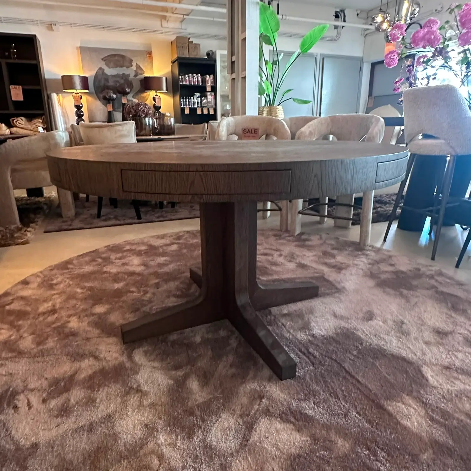 Wide view of a furniture showroom with a large circular wooden table surrounded by beige upholstered chairs on a plush rug.