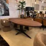 Showroom seating area with an oval wooden table, woven chairs, a tall potted plant, and decorative lamps and sculptures along a sideboard.