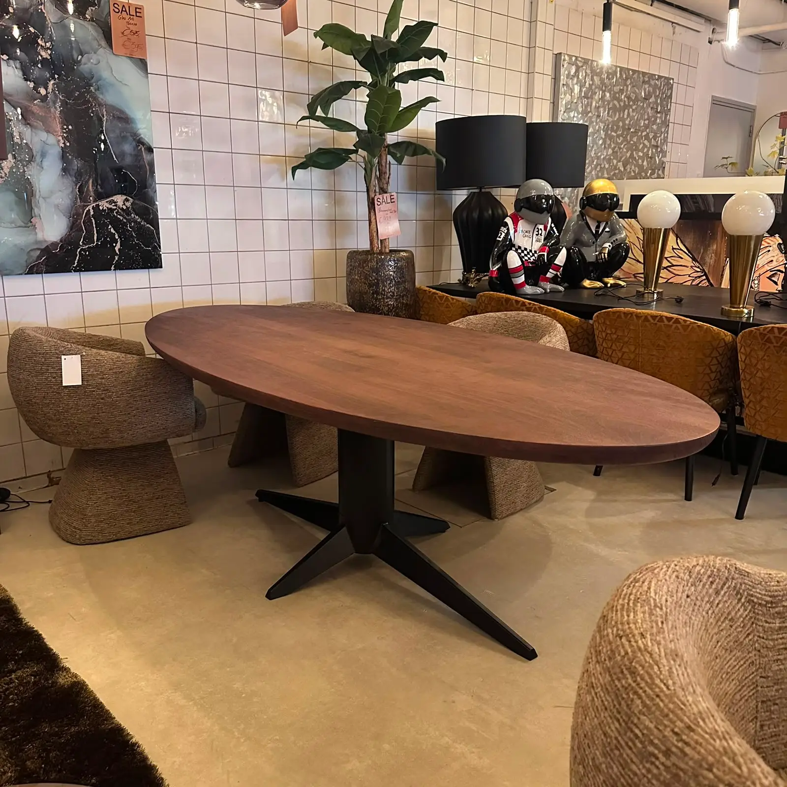 Showroom seating area with an oval wooden table, woven chairs, a tall potted plant, and decorative lamps and sculptures along a sideboard.