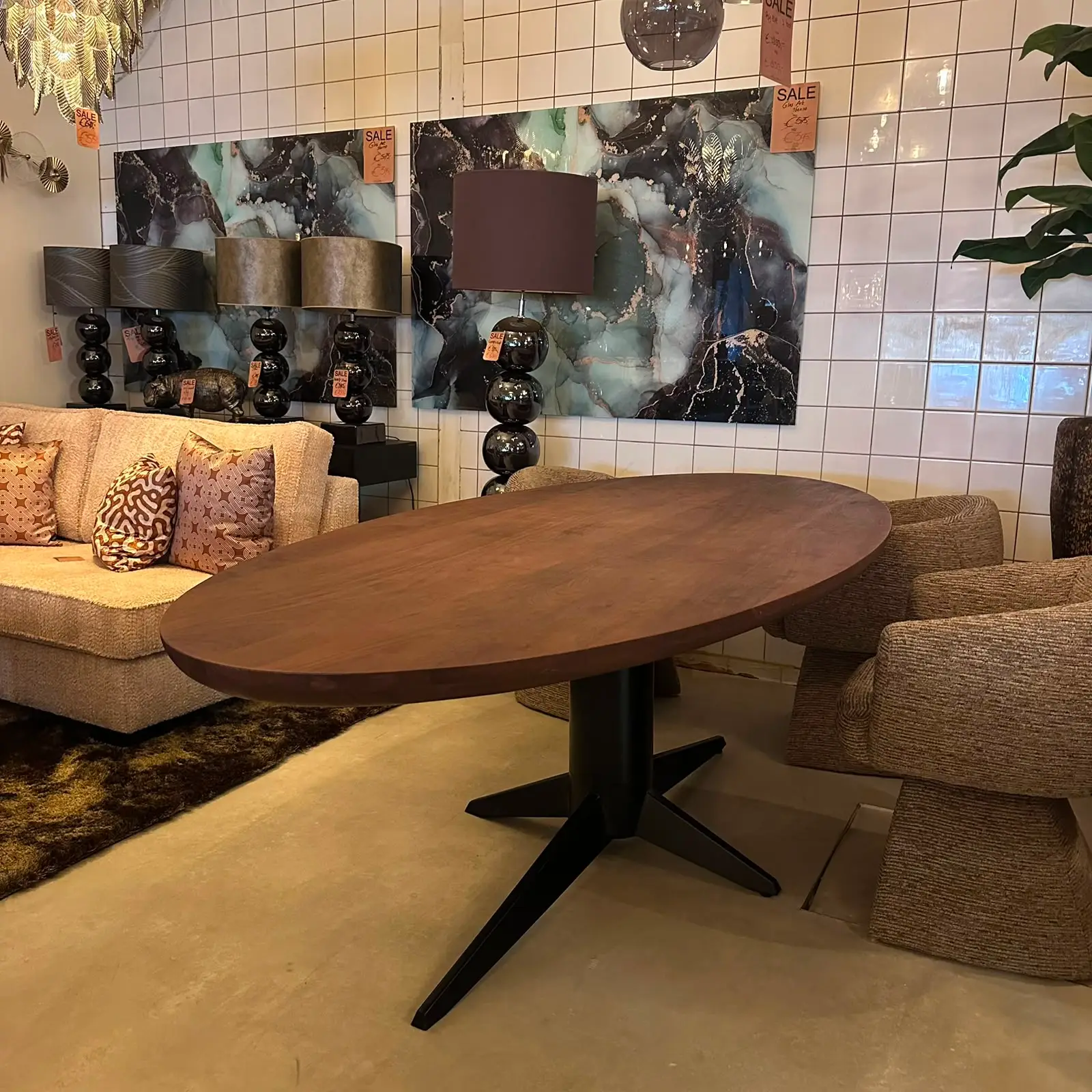 Showroom living area with an oval wooden table, beige sofa with patterned cushions, and black bubble lamps against a white tiled wall with blue marble art panels nearby.